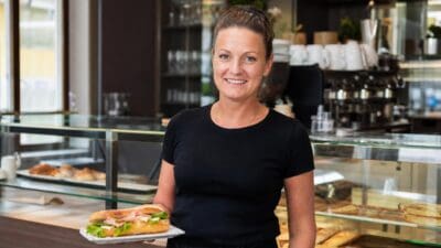 Lächelnde Frau in schwarzem Shirt hält ein belegtes Baguette auf einem Teller