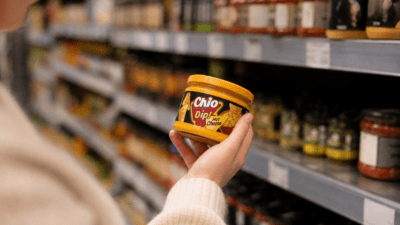 Eine Hand hält ein Glas Chio Dip Hot Cheese vor Supermarktregalen.