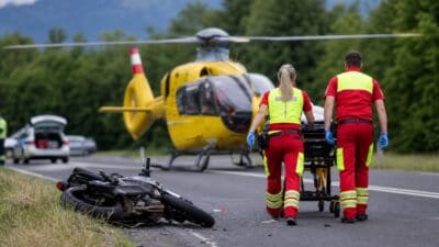 Rettungskräfte schieben eine Trage mit Verletzter zu einem gelben Hubschrauber, Motorrad liegt am Straßenrand