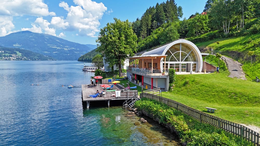 Strandbad mit Holzsteg, bunten Sonnenschirmen und modernem Gebäude am See vor Bergen