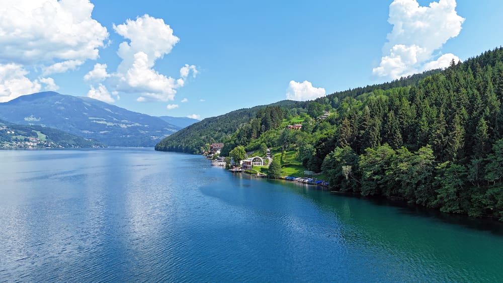 Blick über den blauen See auf bewaldete Hügel mit Gebäuden am Ufer unter blauem Himmel