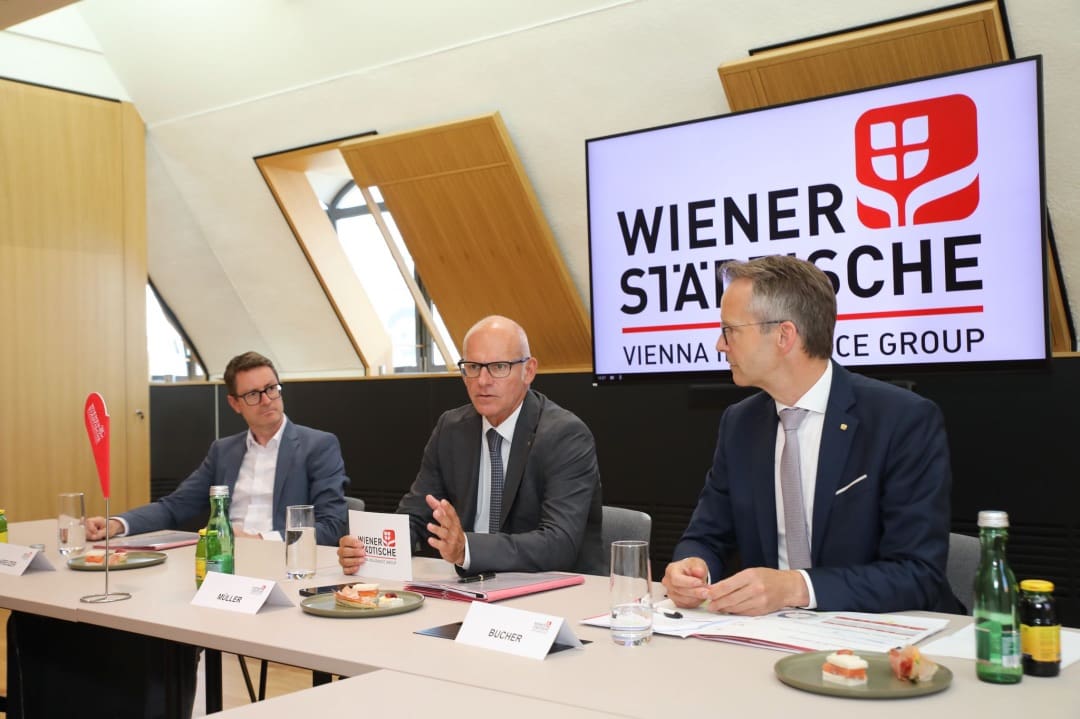 Drei Männer in Anzügen sitzen an einem Konferenztisch vor dem Logo der Wiener Städtischen.