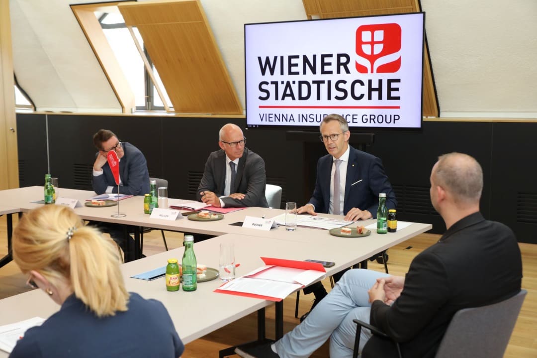 Geschäftsleute sitzen an einem Konferenztisch vor einer Leinwand mit dem Logo der Wiener Städtischen.