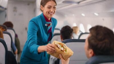 Eine Flugbegleiterin in blauer Uniform reicht einem Passagier ein Döner-Fladenbrot in einem Flugzeug.