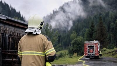 Feuerwehrmann von hinten mit Helm und gelbem Schlauch vor rotem Löschfahrzeug in bewaldeter Berglandschaft
