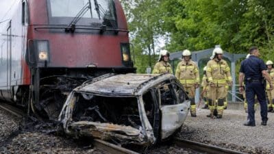Beschädigtes Auto liegt vor rotem Zug auf Bahngleisen mit Feuerwehrleuten und Polizist