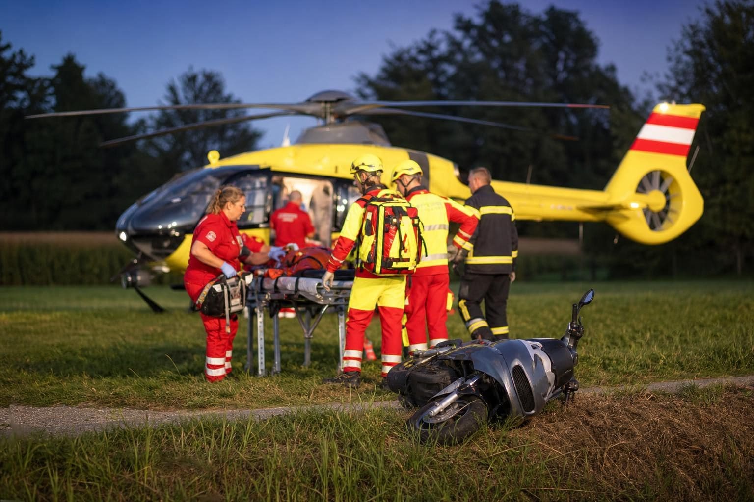 Rettungskräfte mit einem verletzten Patienten vor einem gelben Rettungshubschrauber und einem beschädigten Moped auf einer Wiese
