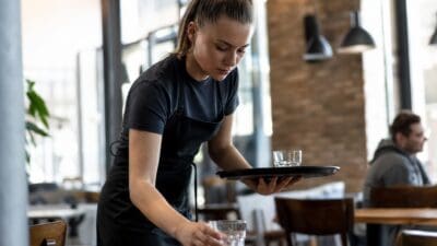 Junge Frau in schwarzer Schürze stellt Gläser auf einen Tisch in einem Restaurant