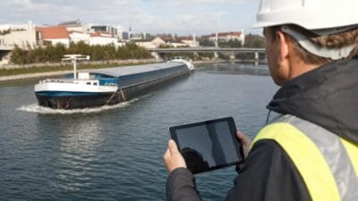 Mann mit Schutzhelm und Sicherheitsweste hält Tablet vor einem Binnenschiff auf dem Fluss
