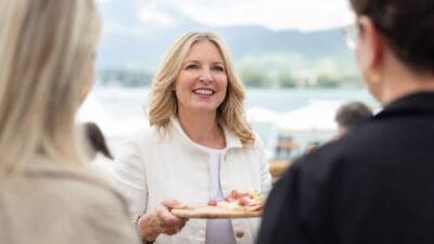 Lachende blonde Frau hält Holzbrett mit Aufschnitt vor dem Wörthersee