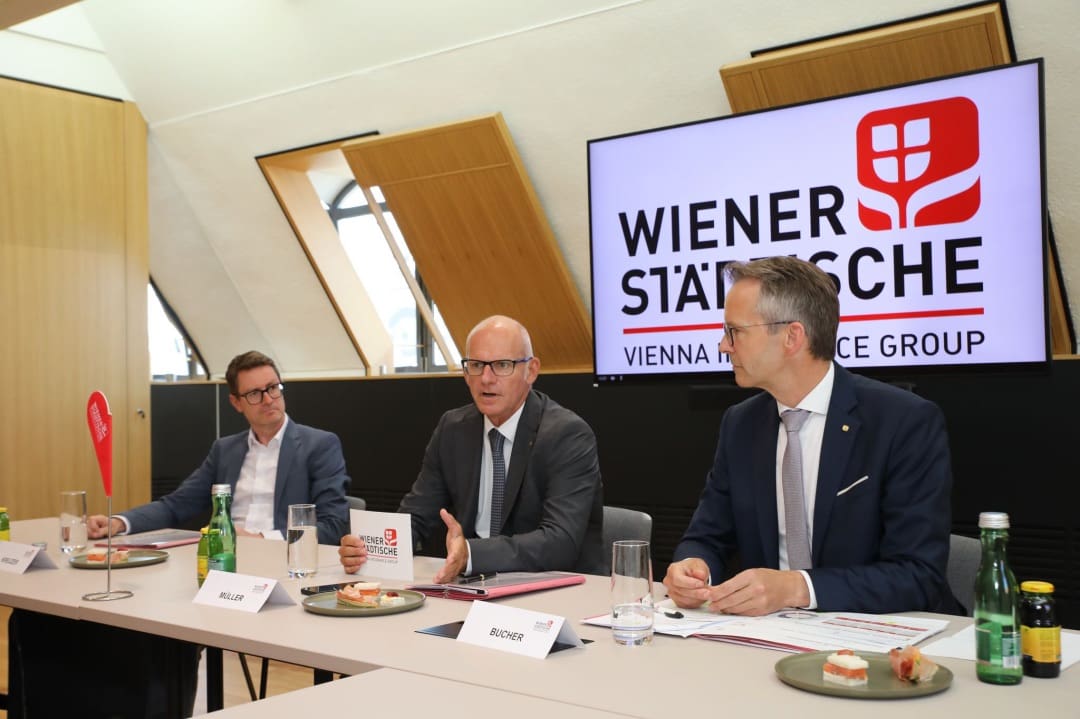Drei Männer in Anzügen sitzen an einem Konferenztisch vor dem Logo der Wiener Städtischen