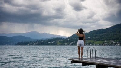 Frau steht auf einem Holzsteg am See unter dunklen Gewitterwolken mit Bergen im Hintergrund