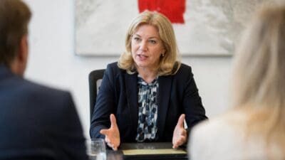 Blonde Frau mit erhobenen Händen spricht an einem Besprechungstisch zu zwei Personen