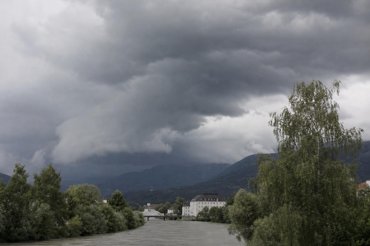 Dunkle Gewitterwolken über einem Fluss mit Gebäuden und Bergen im Hintergrund