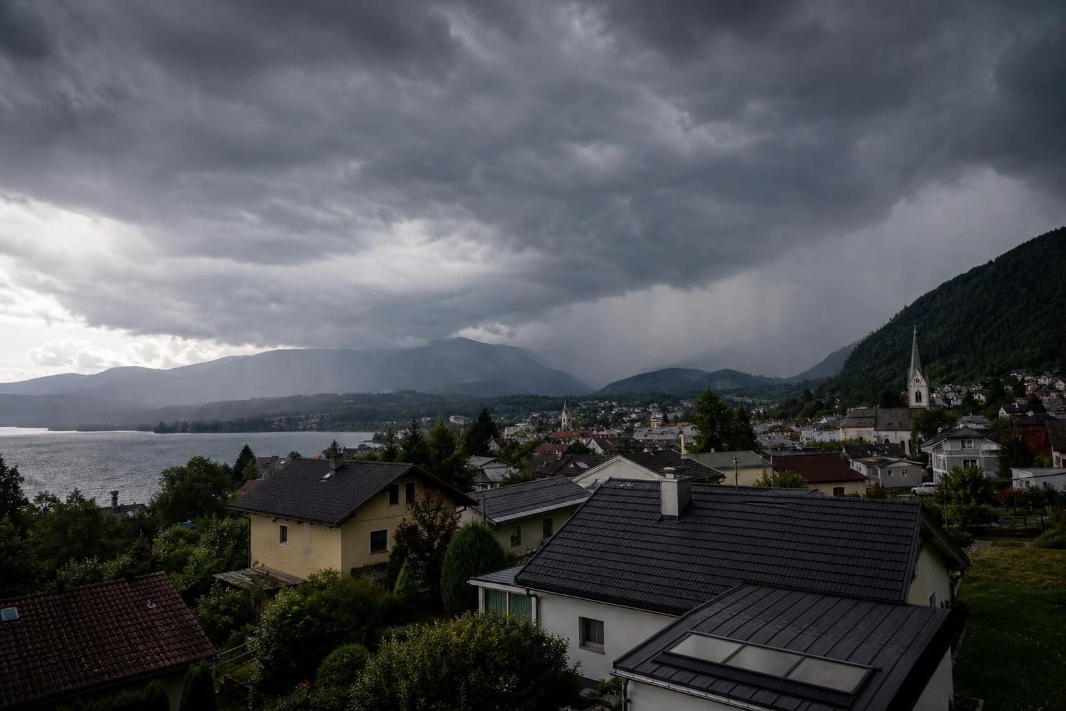 Dunkle Gewitterwolken über einer Ortschaft mit Kirchturm am See und Bergen