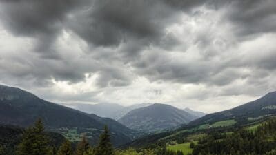 Dunkle Gewitterwolken über einem grünen Bergtal mit Wäldern und Wiesen