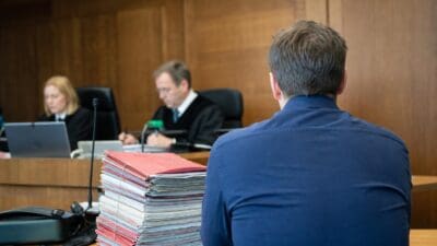 Rückenansicht eines Mannes vor Richter und Beisitzerin im Gerichtssaal mit Aktenstapel