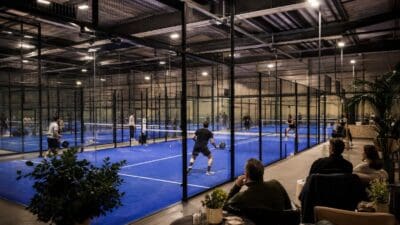 Innenansicht einer großen Padel-Halle mit blauen Courts und aktiven Spielern sowie Zuschauern