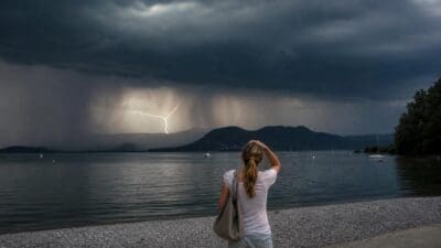 Frau mit Pferdeschwanz steht am Ufer eines Sees und beobachtet Blitze in dunklen Gewitterwolken über dem Wasser
