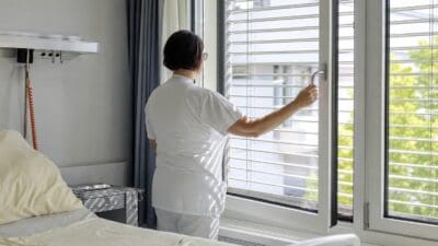 Pflegekraft in weißer Uniform bedient Jalousien an einem Krankenhausfenster neben einem Bett