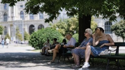 Ältere Frau und Mann sowie junge Frau mit Fächer sitzen auf Parkbänken unter einem Baum.