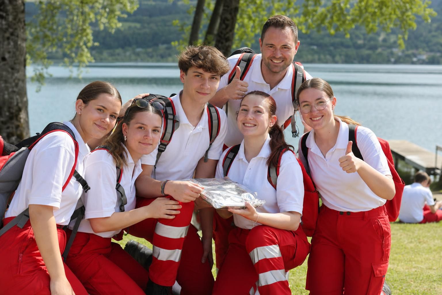Gruppe junger Menschen in roten und weißen Uniformen posiert am Seeufer mit einer Auszeichnung