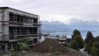 Baustelle eines mehrstöckigen Gebäudes mit Gerüst und Erdhaufen vor See und Bergen