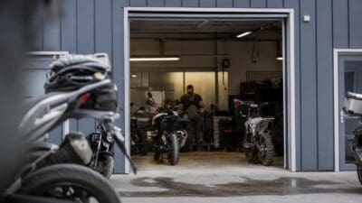 Motorräder vor und in einer blauen Werkstatthalle mit offenem Tor und Mechaniker