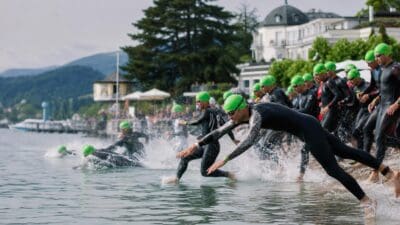 Triathleten in schwarzen Neoprenanzügen und grünen Badekappen laufen ins Wasser des Wörthersees.