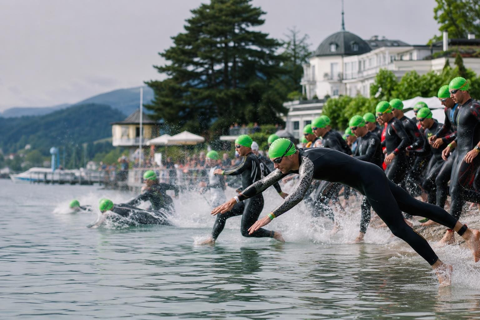 Triathleten in schwarzen Neoprenanzügen und grünen Badekappen laufen ins Wasser des Wörthersees.