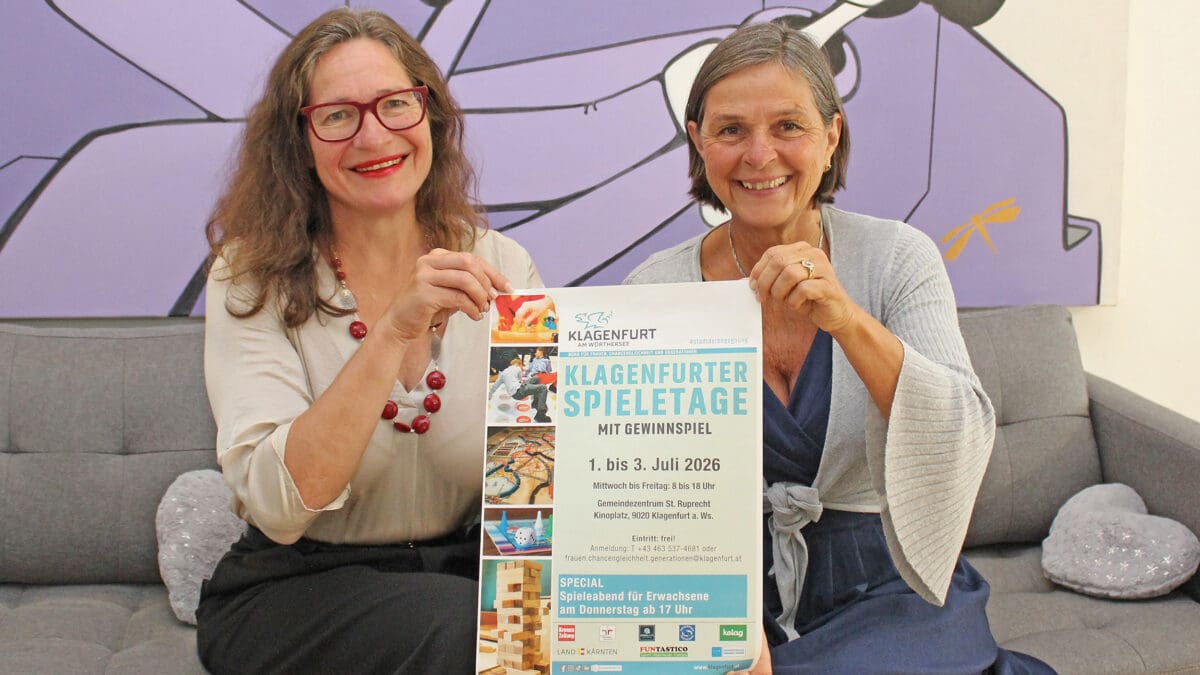 Zwei Frauen halten ein Plakat für die Klagenfurter Spieletage 2026 auf einem Sofa