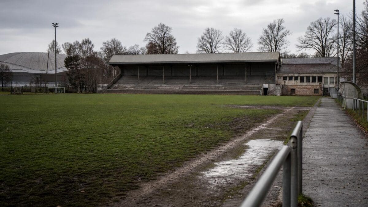 Leerer grüner Fußballplatz mit alter Tribüne und Flutlichtmasten unter bewölktem Himmel