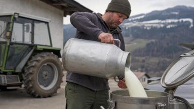 Bauer gießt frische Milch aus einem Aluminiumkanister in einen großen Edelstahltank vor einem Traktor in den Bergen