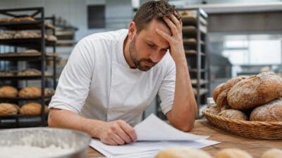 Bäcker in weißer Uniform hält sich den Kopf und betrachtet Papiere in einer Backstube