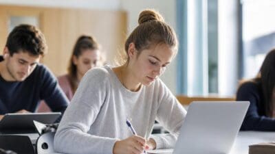 Junge Frau mit hochgestecktem Haar schreibt mit Stift in ein Notizbuch vor einem Laptop in einem Klassenzimmer.