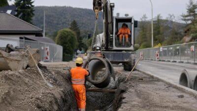 Bauarbeiter in oranger Schutzkleidung positioniert ein großes Betonrohr in einem Graben neben einem Bagger.