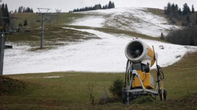 Gelbe Schneekanone auf einer teilweise beschneiten Skipiste mit Sesselliften im Hintergrund