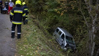 Feuerwehrleute blicken auf einen umgestürzten silbernen Kleinbus in bewaldeter Böschung