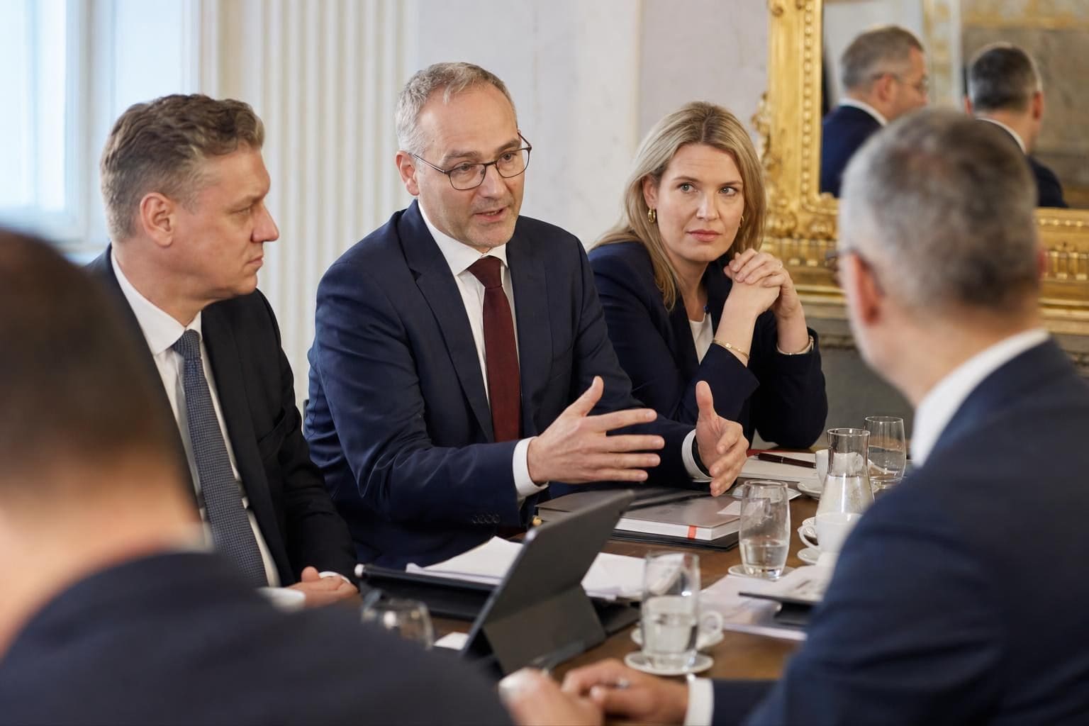 Mehrere Politiker in Anzügen diskutieren an einem Konferenztisch mit Dokumenten und Tablets