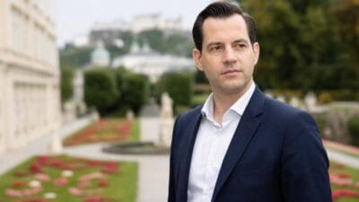 Florian Kreibich im dunklen Anzug steht im Mirabellgarten vor Blumenbeeten und Gebäuden.