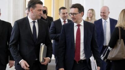 Zwei Männer in dunklen Anzügen gehen nebeneinander durch einen Gang und sprechen miteinander.