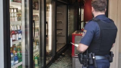 Polizist betrachtet zerbrochene Glasscheiben vor offenen Kühlschränken in einem Kiosk