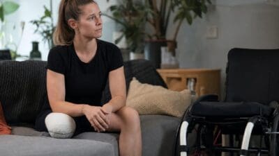 Eine Frau mit bandagiertem Bein sitzt auf einem Sofa neben einem Rollstuhl.