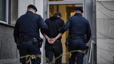 Zwei Polizisten in Uniform führen einen gefesselten Mann in ein Gebäude