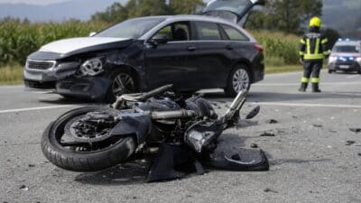 Beschädigtes Motorrad liegt auf der Straße vor einem verunfallten schwarzen Pkw mit offenem Kofferraum.