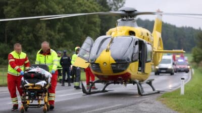 Rettungskräfte schieben eine Trage mit einem Patienten zu einem gelben Rettungshubschrauber auf einer Straße.