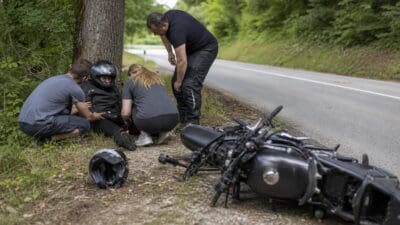 Helfer versorgen eine sitzende Motorradfahrerin mit Helm am Straßenrand neben einem umgestürzten Motorrad