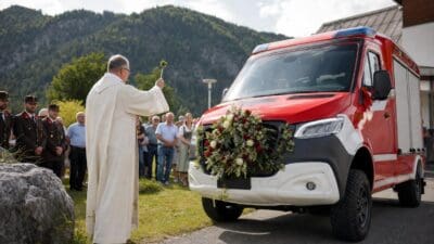 Ein Priester segnet ein mit Blumen geschmücktes rotes Feuerwehrfahrzeug vor einer Menschenmenge.