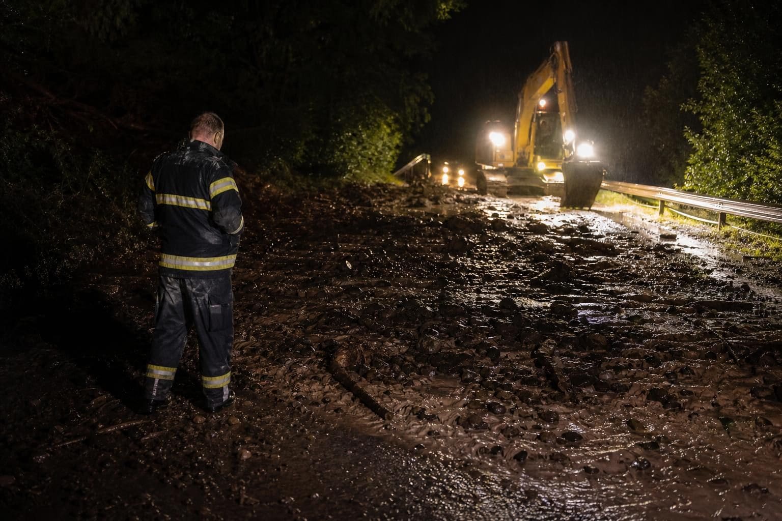 Feuerwehrmann in Uniform steht nachts auf schlammbedeckter Straße vor einem Bagger
