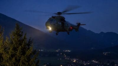Ein grüner Militärhubschrauber fliegt bei Nacht über einem beleuchteten Tal mit Bergen im Hintergrund